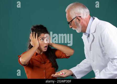 Erfahrener Arzt, der jungen Patienten, die Pillen ablehnen und die Hände an den Ohren halten, eine Handfläche voller Medikamente anbietet. Frau Angst vor Gesundheit Auto Stockfoto