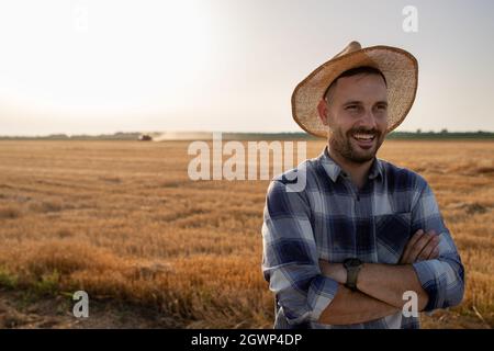 Porträt eines Bauern mit gekreuzten Armen, der Strohhut trägt und im Sommer auf dem geernteten Feld steht Stockfoto