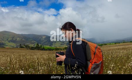 Wunderschöne hispanische Forscherin, von hinten gesehen, mit Rucksack, der Fotos auf ihrer Kamera an einem bewölkten Morgen inmitten eines gesät Feldes ansieht Stockfoto