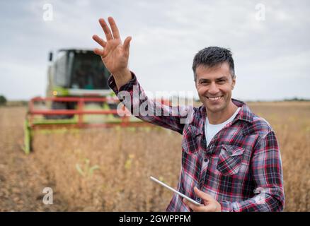 Männlicher Agronom lächelt, winkt mit der Hand und verwendet moderne Tablet-Technologie in der Landwirtschaft. Mann, der im Sojafeld steht und im Hintergrund die Erntemaschine arbeitet Stockfoto