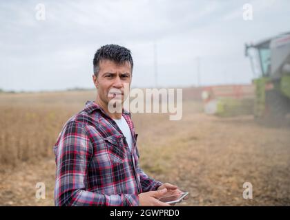 Männlicher Agronom lächelt und verwendet moderne Tablet-Technologie in der Landwirtschaft. Mann, der im Sojafeld steht und im Hintergrund die Erntemaschine arbeitet. Stockfoto