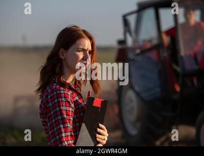 Hübsche Agronomin, die im Feld steht und Notizen mit Clipboard gemacht hat. Bauer sitzt im Traktor mit geöffneter Tür im Hintergrund. Stockfoto