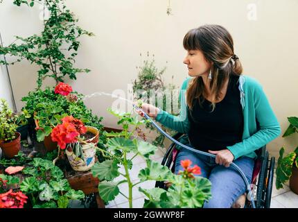 Junge Frau im Rollstuhl, die Pflanzen im Hinterhof ihres Hauses wässern Stockfoto