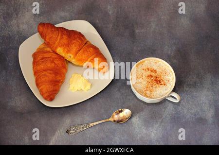 Eine Tasse Cappuccino und zwei frische Croissants mit Butter auf einer weißen Keramikplatte. Blick von oben. Stockfoto