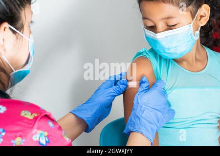 Arzt in blauen Gummischutzhandschuhen, die einem Mädchen einen klebenden Verband auf den Arm legen, nachdem sie ihre Haut verletzt oder einen Impfstoff injiziert hat. Erste Hilfe. Medizinisch, p Stockfoto