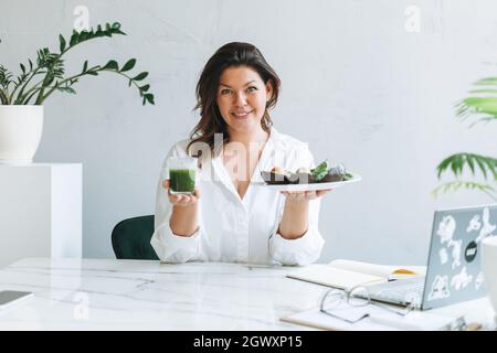 Junge lächelnde Brünette Frau Ernährungsberater Arzt plus Größe in weißem Hemd mit grünen gesunden Lebensmitteln im modernen hellen Bürozimmer. Arzt kommuniziert Stockfoto