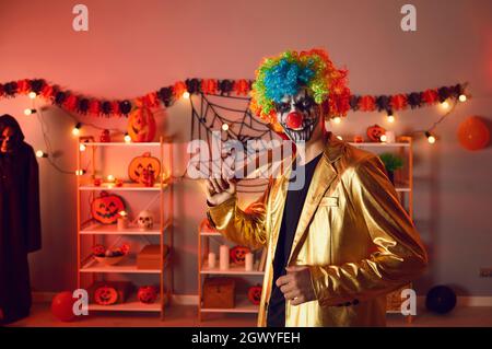 Portrait eines erwachsenen Mannes, der als gruseliger böser Clown mit Axt auf der Halloween Party verkleidet ist Stockfoto