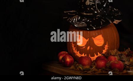 Geschnitzter orangefarbener Kürbis auf dunklem Hintergrund. Rote Äpfel und einige trockene Blätter liegen auf einem Holztisch. Jack-o-Laterne mit schwarzem Hut, der Stimmung für Trick-o setzt Stockfoto
