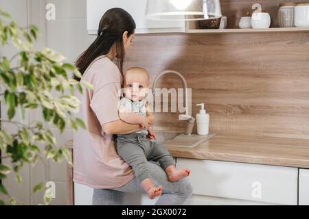 Beschäftigt Mutter Reinigung Küche mit Baby auf den Armen. Frau kocht und hält Säugling, müde Frau macht tägliche Hausaufgaben mit Kind. Stockfoto