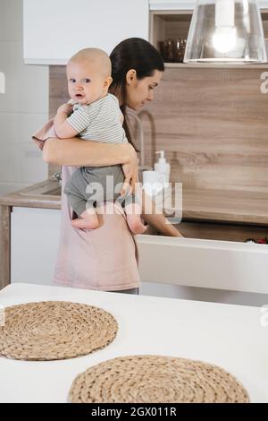 Beschäftigt Mutter Reinigung Küche mit Baby auf den Armen. Frau kocht und hält Säugling, müde Frau macht tägliche Hausaufgaben mit Kind. Stockfoto