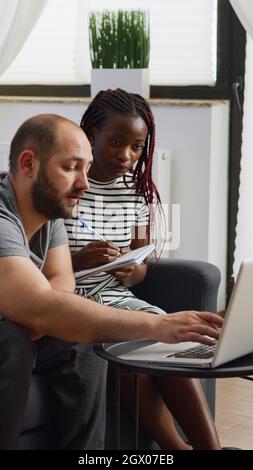 Junges interrassisches Paar, das mit einem Laptop und einem Notebook im Wohnzimmer ein Budget für Steuerzahlungen plant. Multiethnische Menschen berechnen Geld für Steuern. Mann und Frau überprüfen die Buchhaltung Stockfoto