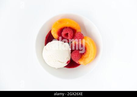 Kugel hausgemachtes Vanilleeis in einer weißen Schüssel mit pfirsichfarbenen Hälften, frischen Himbeeren und Himbeersoße auf Weiß isoliert. Stockfoto