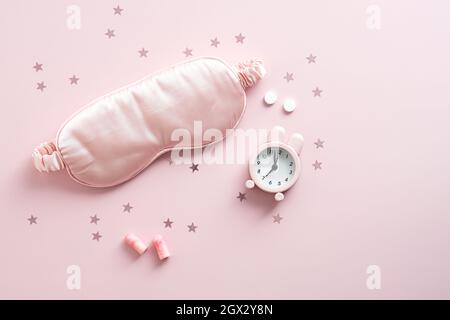 Flache Schlafmaske, Wecker, Ohrstöpsel und Pillen auf rosa Hintergrund. Zubehör für besseren Schlaf in der Nacht Stockfoto