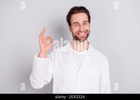 Foto von fröhlichen ziemlich junge Kerl Arzt Arbeitskleidung zeigt okey Zeichen lächelnd isoliert grauen Hintergrund Stockfoto