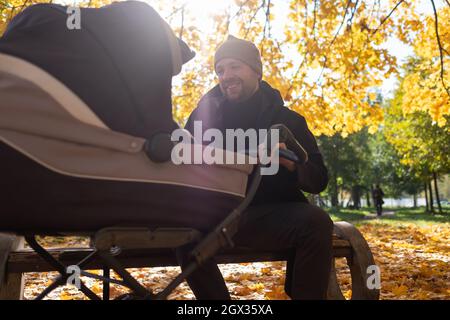 Glücklicher junger Vater mit Kinderwagen sitzt auf der Bank in der Natur im Park Stockfoto