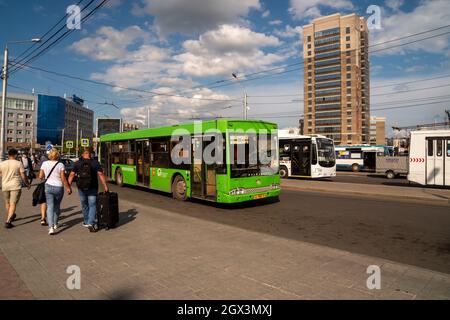 Menschen mit einem Koffer eilen an einem sonnigen Sommertag zu einem Stadtbus, der an einer Bushaltestelle auf dem Bahnhofsplatz geparkt ist. Stockfoto