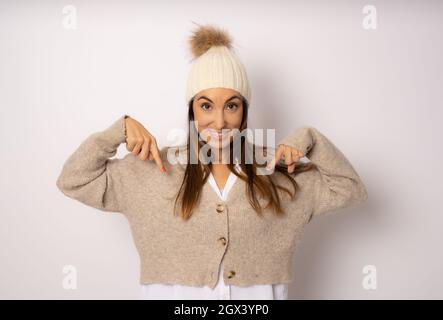 Die junge, schöne Frau, die einen legeren Pullover und einen Wollhut trägt, überraschte und zeigte mit den Fingern und erhobenen Armen nach unten Stockfoto
