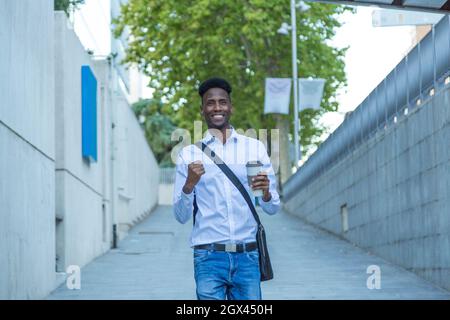 Porträt eines jungen afroamerikanischen Geschäftsmannes mit einem Glas Kaffee in der Hand und triumftiger Ausdruck, der die Straße entlang geht Stockfoto