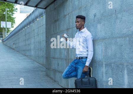 Junger schwarzer Geschäftsmann, der sich mit einem Glas Kaffee und einer Aktentasche an der Wand lehnte Stockfoto