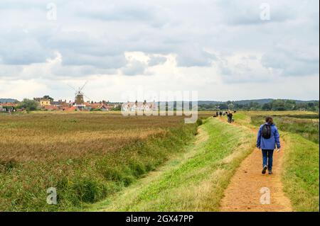 Menschen, die auf dem Blakeney Freshes Rundwanderweg mit Blick auf das Dorf Cley on the Sea mit seiner Windmühle, Norfolk, England, wandern. Stockfoto