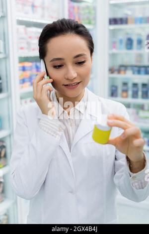 Fröhlicher Apotheker im weißen Mantel, der auf dem Smartphone spricht und die Flasche in der Drogerie hält Stockfoto