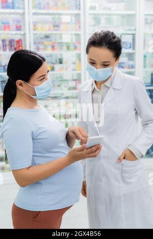 asiatische Schwangerin einer medizinischen Maske, die auf das Smartphone in der Nähe des Apothekers in der Drogerie zeigt Stockfoto