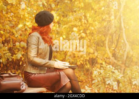 Herbstporträt. Hübsche Frau mit grafischem Tablet auf Parkbank. Rothaarige Mädchen in Baskenmütze und modische Jacke sieht weg. Lederrucksack auf einer Holzbank. Designer oder Freiberufler. Stockfoto