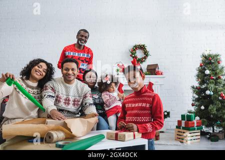 Glückliche afroamerikanische Familie, die die Kamera anschaut, während sie Geschenke im Wohnzimmer mit weihnachtsdekoration verpackt Stockfoto