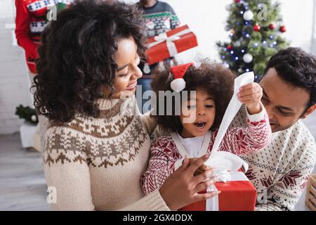 Erstaunt afroamerikanische Mädchen Eröffnung weihnachtsgeschenk in der Nähe glückliche Eltern und verschwommen weihnachtsbaum Stockfoto