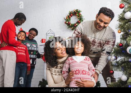 Fröhliche afroamerikanische Eltern umarmen Tochter in der Nähe verschwommen Sohn mit Großeltern im Wohnzimmer mit weihnachtsdekoration Stockfoto