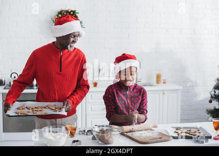 Glücklicher afroamerikanischer Mann, der in der Nähe des Enkels Backblech mit weihnachtsgebäck hält Stockfoto