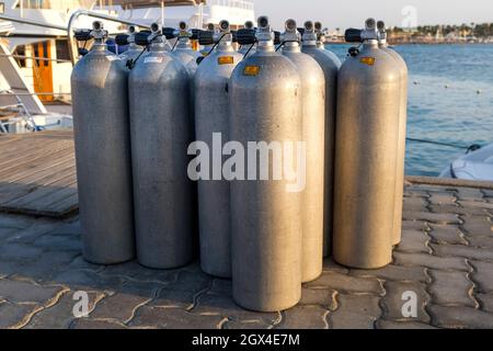 Viele Alu-Tauchsauerstofftanks stehen vor den grünen Strandbüschen und Felsen auf dem Sand in Thailand mit Kopierraum Stockfoto