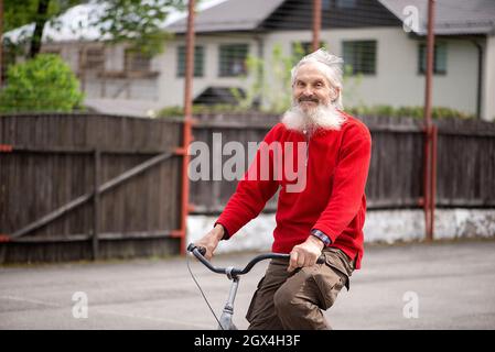 Positiv lächelnder älterer bärtiger Mann fährt mit einem roten Pullover auf dem Fahrrad, die Haare fließen im Wind Stockfoto