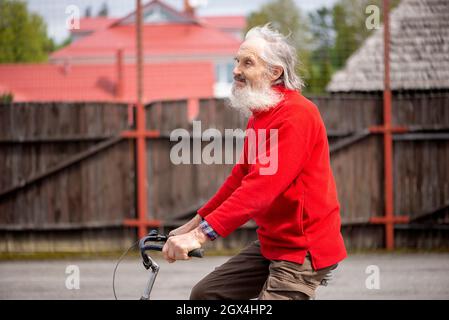Trauriger alter bärtiger Mann, der mit einem roten Pullover auf einem Fahrrad fährt, Haare flattern im Wind Stockfoto
