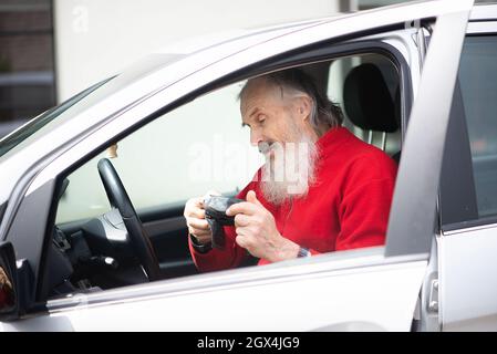 Ein älterer Mann mit grauem Bart sitzt im Auto und hält eine Fotokamera Stockfoto