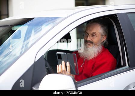Bärtiger älterer Mann mit Brillen, die das Telefon halten und SMS schreiben, während er auf dem Parkplatz im Auto sitzt Stockfoto