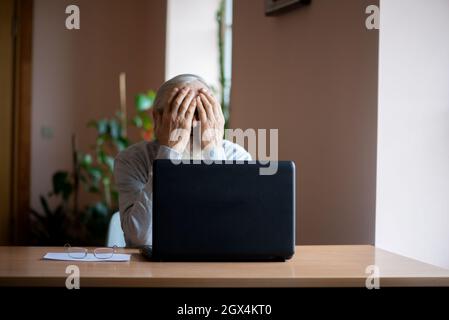 Ein müder, gebrochener, resigniert, beschäftigt älterer Mann sitzt vor einem Laptop, Computer und bedeckt sein Gesicht mit der Hand. Der traurige Senior überprüft seine Klimaanlage Stockfoto