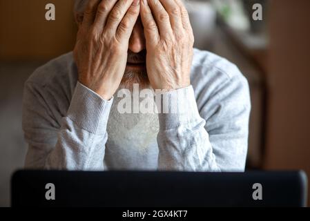 Ein müder, gebrochener, resigniert, beschäftigt älterer Mann sitzt vor einem Laptop, Computer und bedeckt sein Gesicht mit der Hand. Der traurige Senior überprüft seine Klimaanlage Stockfoto