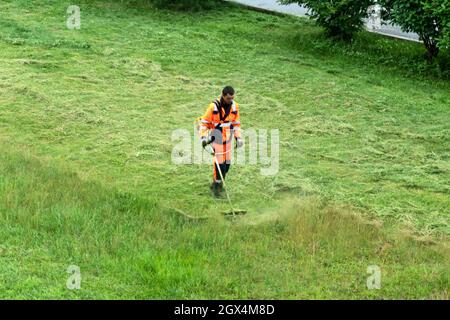 Ein Arbeiter in orangefarbenen Overalls mäht an einem bewölkten Sommertag den Rasen. Stockfoto