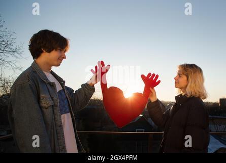 Junger Mann und ein verliebtes Teenager-Mädchen vor dem Hintergrund des Sonnenuntergangs. Sie stehen einander gegenüber, halten ein großes weiches Herz fest, nehmen Augenkontakt auf, lächeln. L Stockfoto