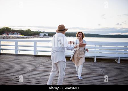 Glückliches Seniorenpaar, das verliebt im Freien auf dem Pier am Meer tanzt und sich anschaut. Stockfoto