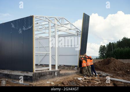 Im Sommer installieren Arbeiter auf einer Baustelle mit den Händen eine Wandtafel am Rahmen eines Industriegebäudes. Stockfoto