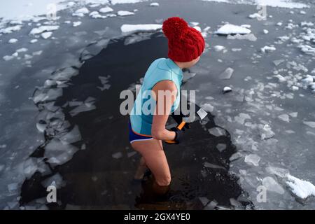 High-Angle-Ansicht einer aktiven älteren Frau im Badeanzug, die im Winter mit Axt Eis bricht, kalt-Therapie-Konzept. Stockfoto