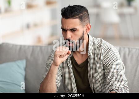 Coronavirus, Influenza-Konzept. Kranker junger arabischer Mann, der in die Faust hustet, auf der Couch sitzt und sich zu Hause unwohl fühlt Stockfoto