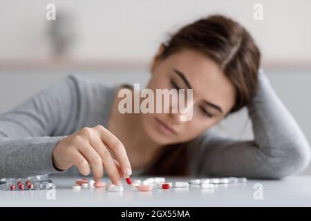 Gleichgültige kaukasische junge Frau, die an Depressionen leidet und viele verstreute Pillen auf dem Tisch berührt Stockfoto