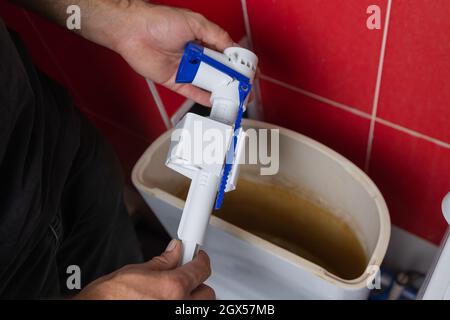 Austausch von Teilen des Toilettentanks. Ein Mann in orangefarbenen Handschuhen repariert den Abfluss des Toilettenbehälters Stockfoto