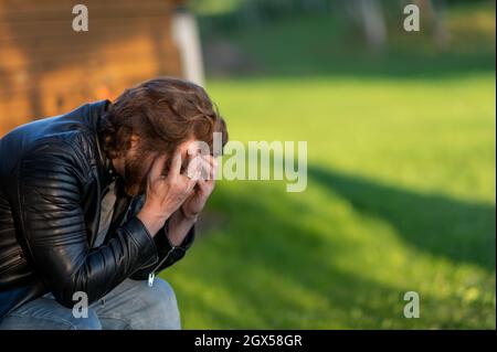 Ein trauriger junger Mann, der sein Gesicht in seinen Handflächen versteckt, sitzt und denkt nach draußen Stockfoto