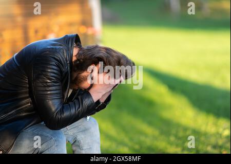 Ein trauriger junger Mann, der weinend sein Gesicht in seinen Handflächen versteckt, sitzt und denkt draußen nach Stockfoto