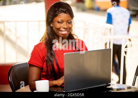 Schöne junge afrikanische Frau, die unter freiem Himmel in der Nähe eines Restaurants sitzt und einen Laptop benutzt, während sie ein Lächeln in der Hand hält Stockfoto