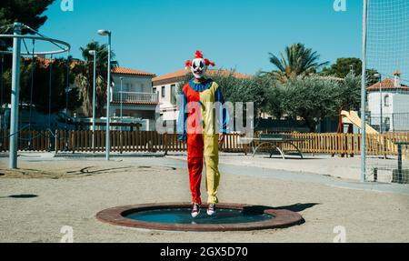 Ein gruseliger Clown, der ein farbenfrohes Gelb-, Rot- und Blaukostüm trägt, hüpft auf einem Trampolin auf einem öffentlichen Spielplatz im Freien Stockfoto